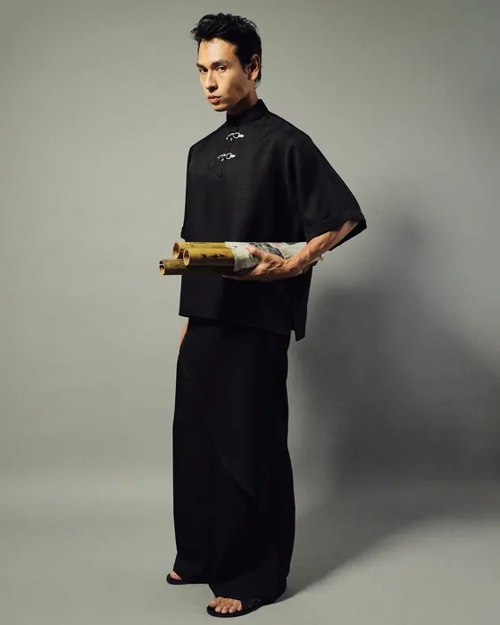 Model in the RIROYO set in black, two silver fireman clasps at the collar, against a grey studio wall