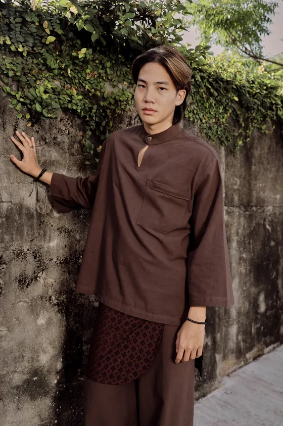 Model in the THUNDUK Tanah set, hand on a stained concrete wall under ivy, the embroidered samping at the waist